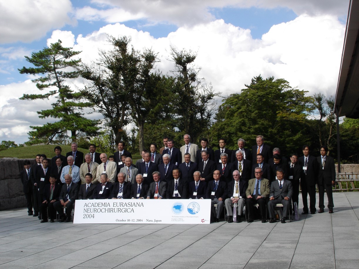Kilkadziesiąt os&oacute;b ubranych w elegancko w garnitury, większość ma zawieszone na szyi plakietki o nieczytelnej treści. Osoby są ustawione w trzech rzędach. Przed pierwszym rzędem stoi duży baner z napisem Academia Eurasiana Neurochirurgica 2024 October 10-12, 2004, Nara Japan